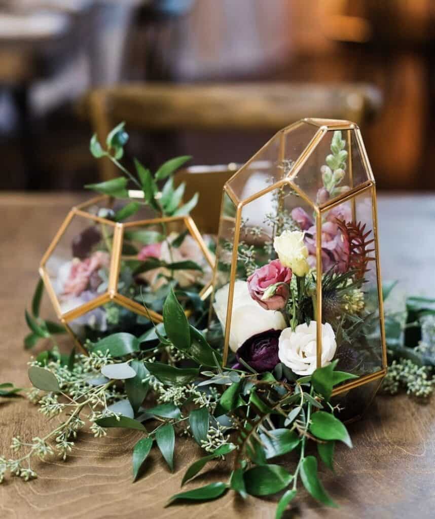 Vase with mixed flowers and greenery inside geometric glass terrarium centerpiece.