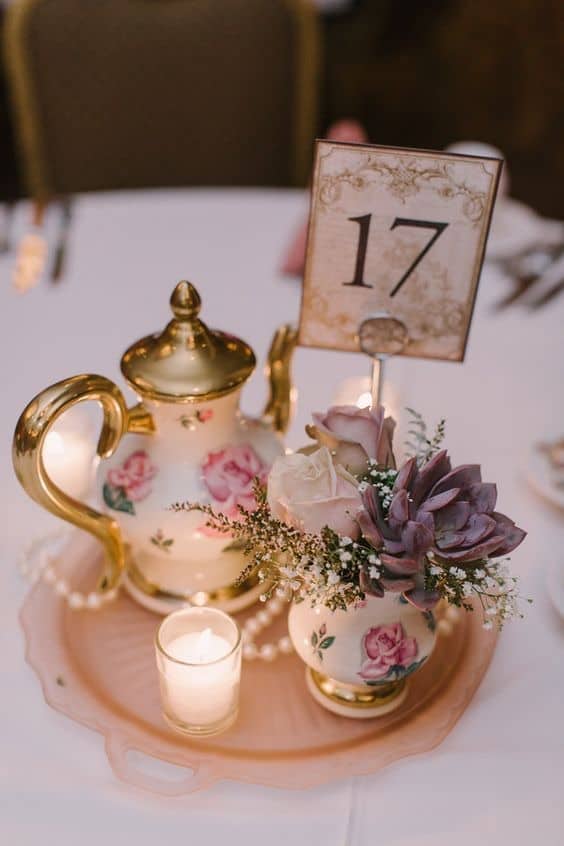 Elegant floral table centerpiece with vintage teapot, candle, and table number card.