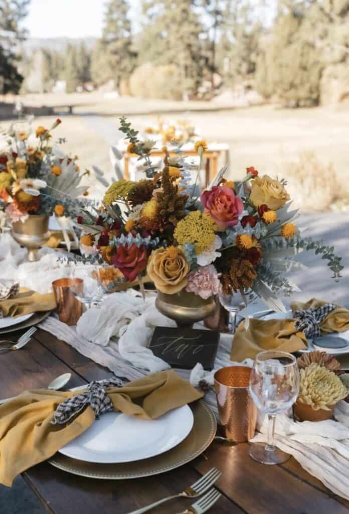 Vibrant autumn floral centerpiece at outdoor wedding reception table with colorful roses and greenery.