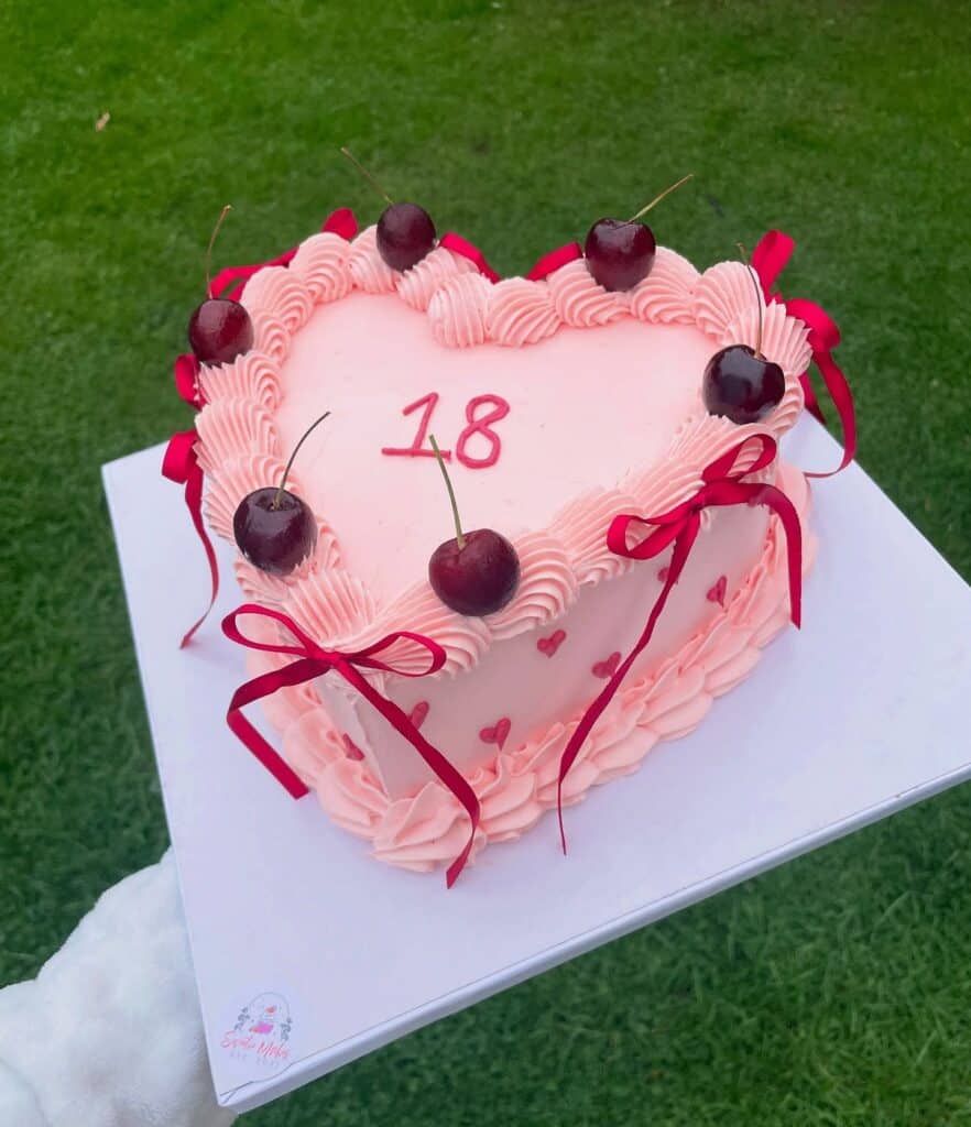 Heart-shaped pink birthday cake with cherries and red ribbons, perfect for 18th celebration.