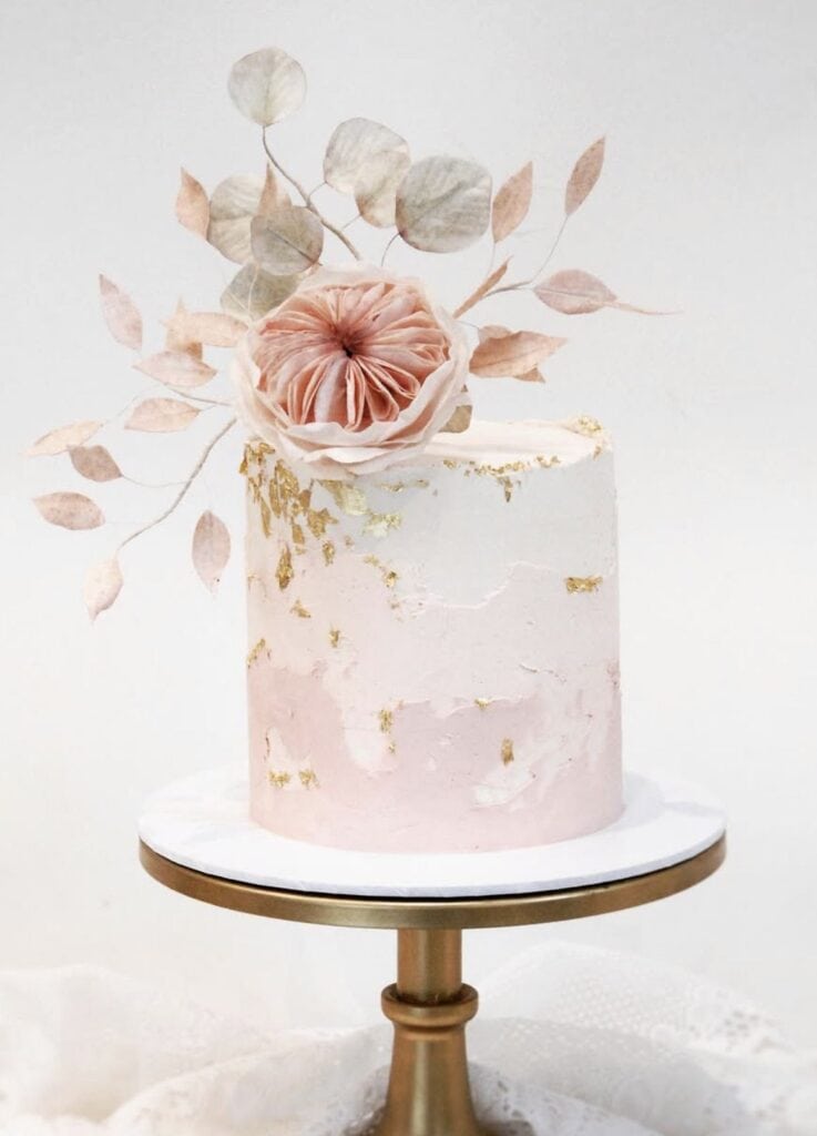 Elegant pink and gold wedding cake with floral topper on gold stand.