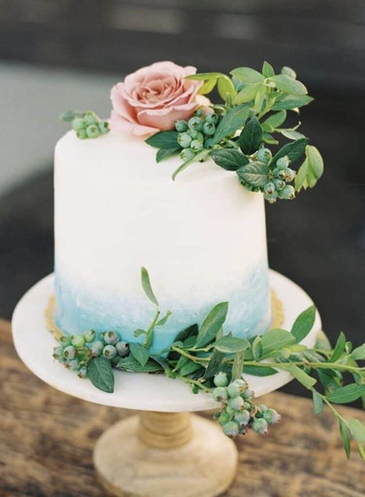 Elegant white wedding cake with blush pink rose and greenery decoration on wooden stand, celebration cake.