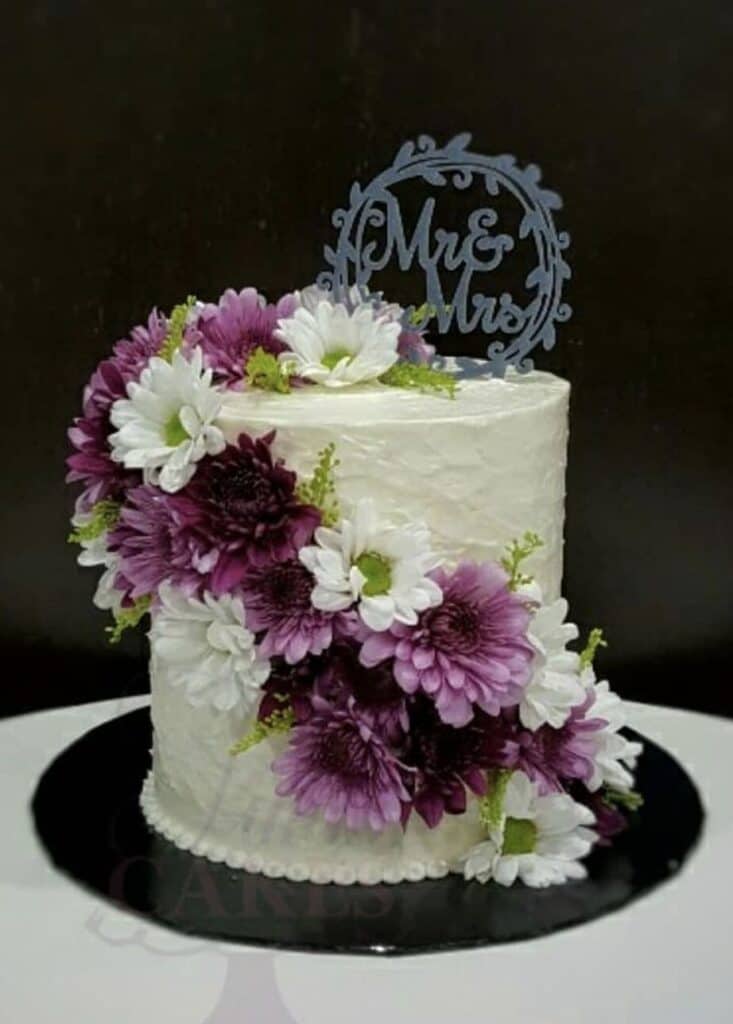 Elegant wedding cake with white frosting and purple and white floral decorations, "Mrs. & Mr." topper, perfect for wedding celebrations.