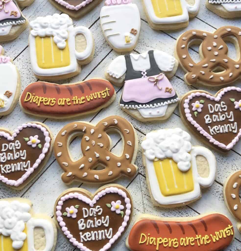 Colorful decorated baby shower cookies with beer mugs, pretzels, and themed messages on a white wooden background.