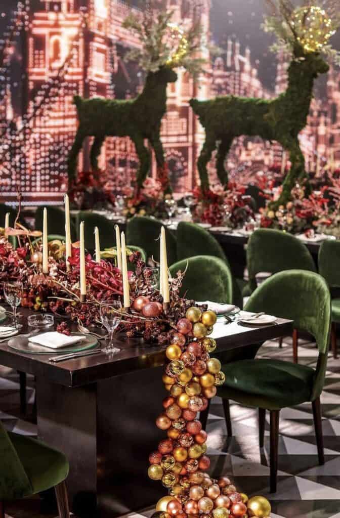 Festive holiday table decor with gold and red ornaments, candles, and reindeer topiary.