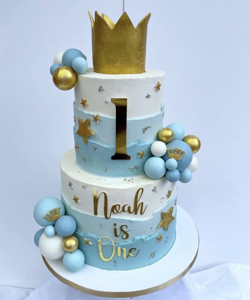 Gold crown topper with blue and white balloon accents on a pastel blue and white first birthday cake.