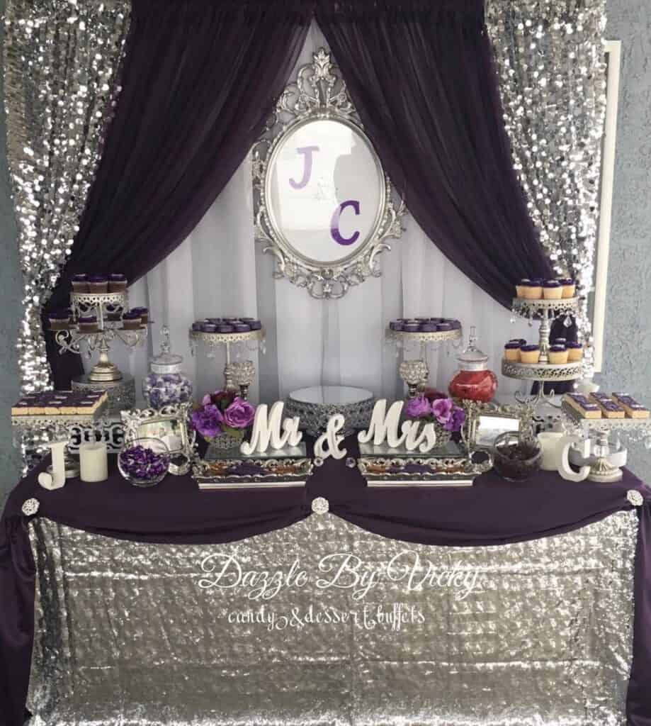 Elegant purple and silver wedding dessert table with floral accents and monogram mirror.