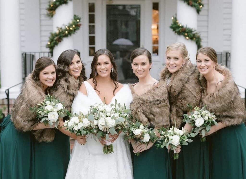 Elegant wedding bridal party with fur shawls and white bouquets outside a historic house.