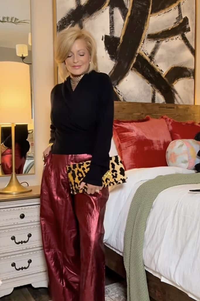Elegant woman in black top and red satin pants, holding leopard print clutch, in stylish bedroom setting.