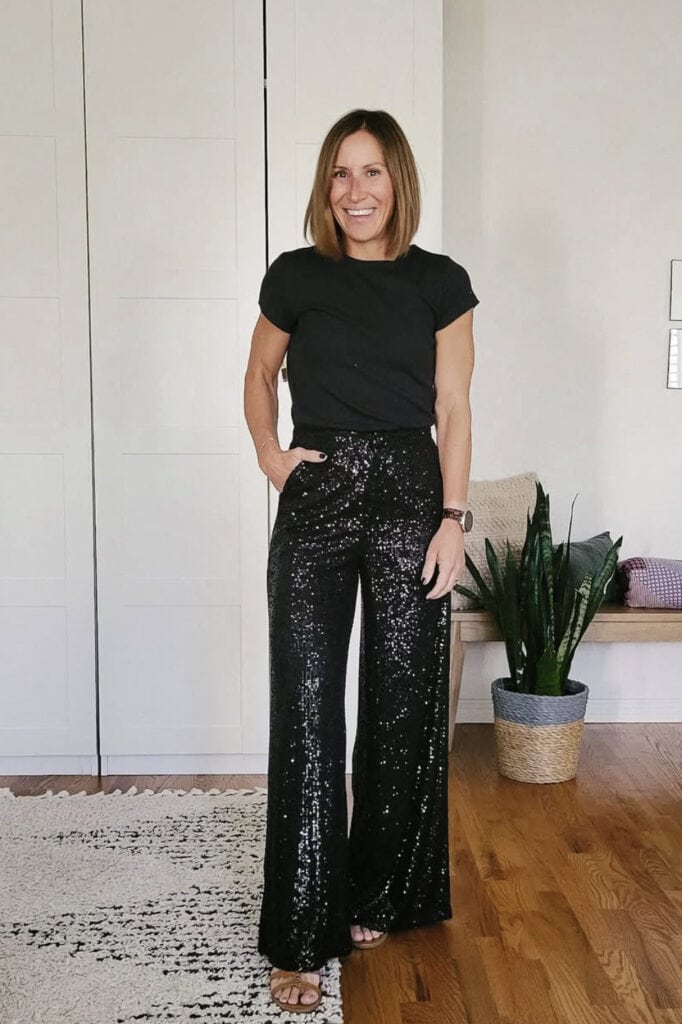 Sh woman wearing black sequin pants and T-shirt standing indoors with houseplant.