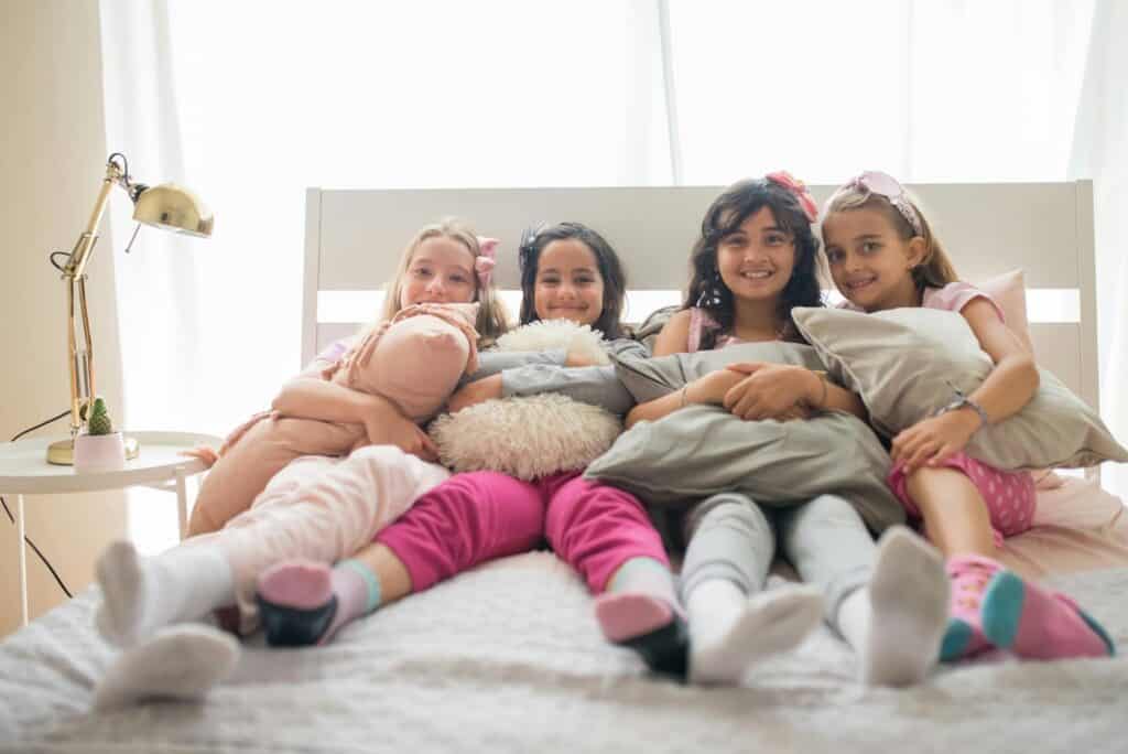 Four cheerful girls enjoying a cozy sleepover party on a bed with pillows and pajamas.