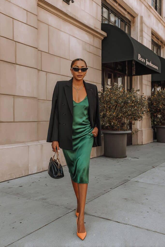 Elegant woman in green satin slip dress with black blazer and accessories, city street fashion.