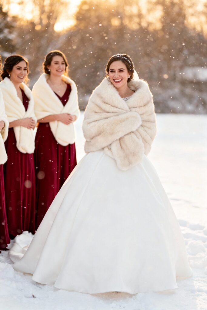 Elegant winter bride in white gown with fur shawl and bridesmaids in burgundy dresses, snowy outdoor wedding.