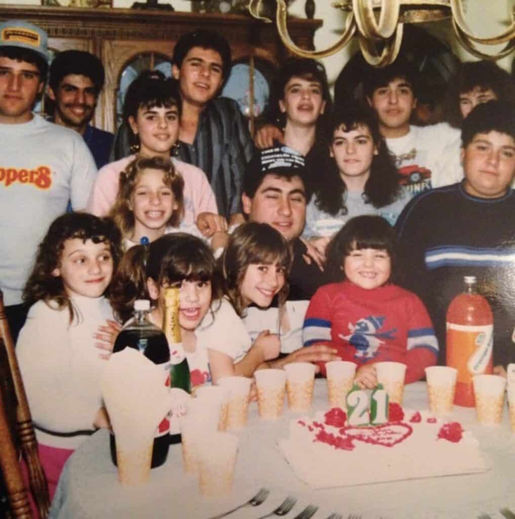 A group of children and adults celebrating a 21st birthday party, with a cake and party drinks.