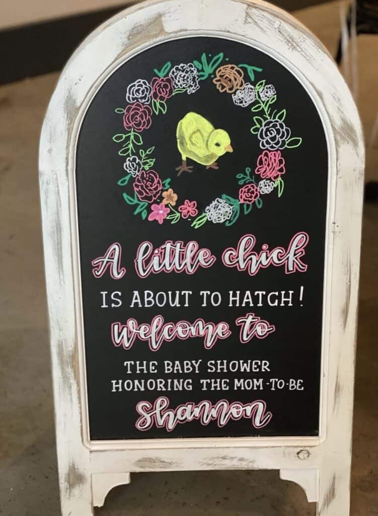 Bright chalkboard sign for a baby shower with floral and chick illustrations, personalized message.