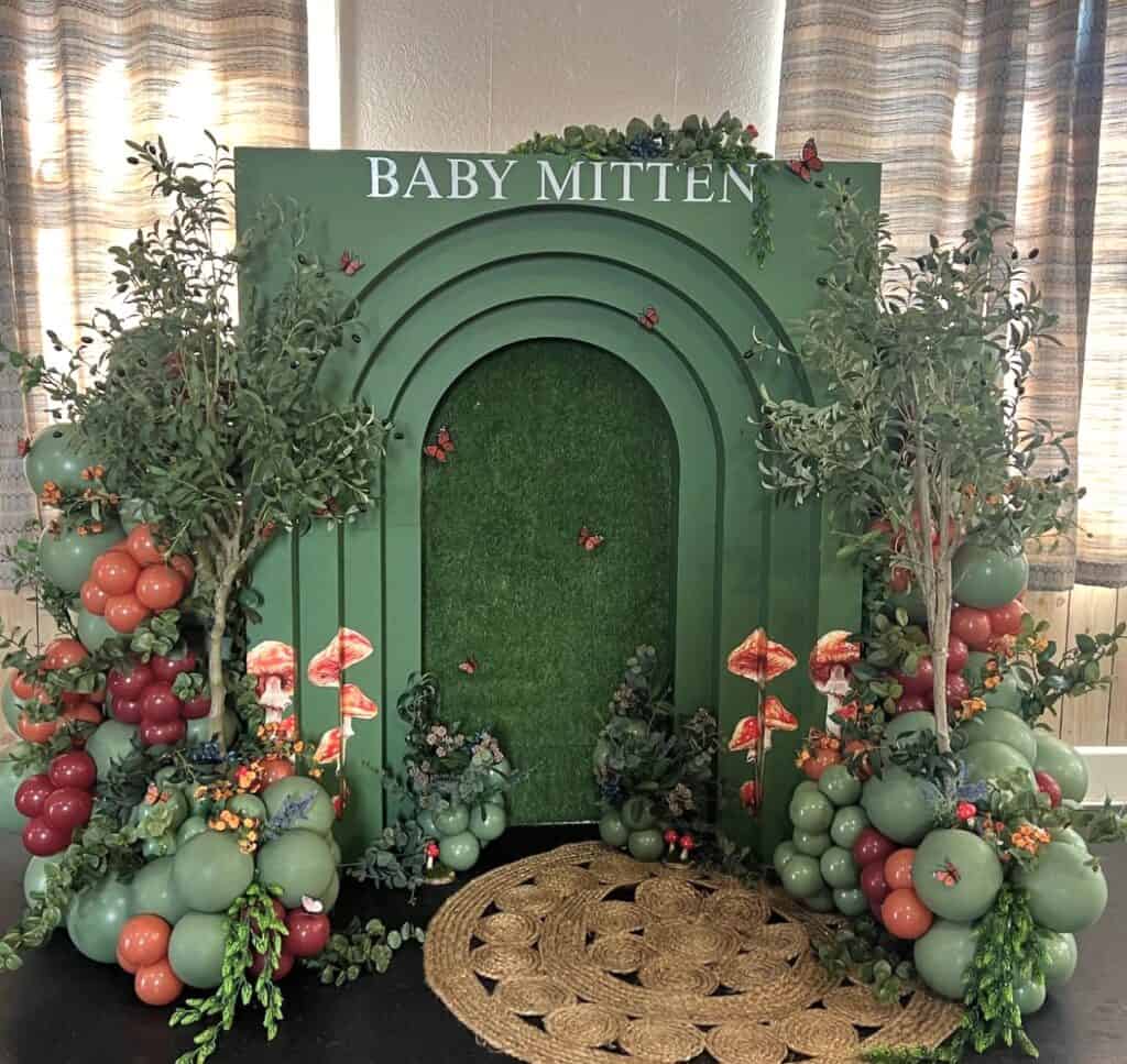 Vibrant green baby shower backdrop with balloons, greenery, mushrooms, and butterflies.