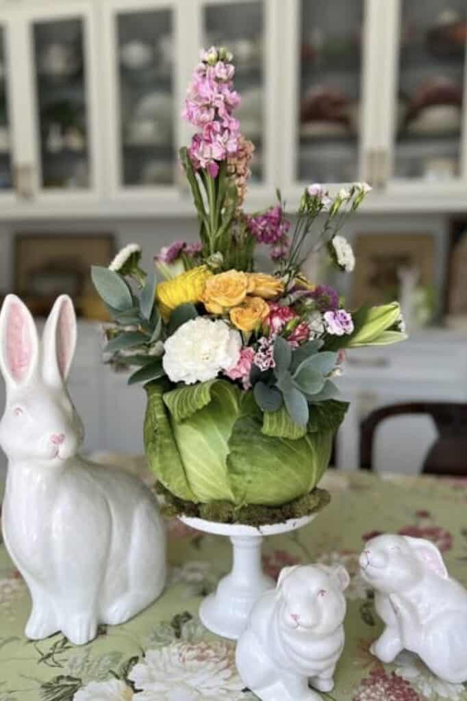Colorful spring flower arrangement in a cabbage boat with ceramic bunny and bunny figurines.