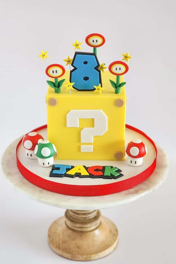 A colorful Super Mario-themed birthday cake with question mark block, mushrooms, and star decoration.