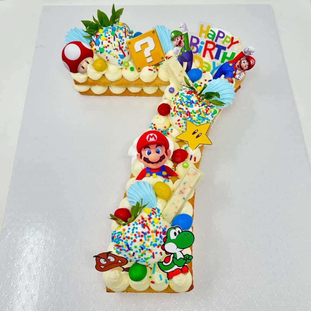 Super Mario-themed birthday cake shaped as the number 7 with Mario, Yoshi, and colorful decorations.
