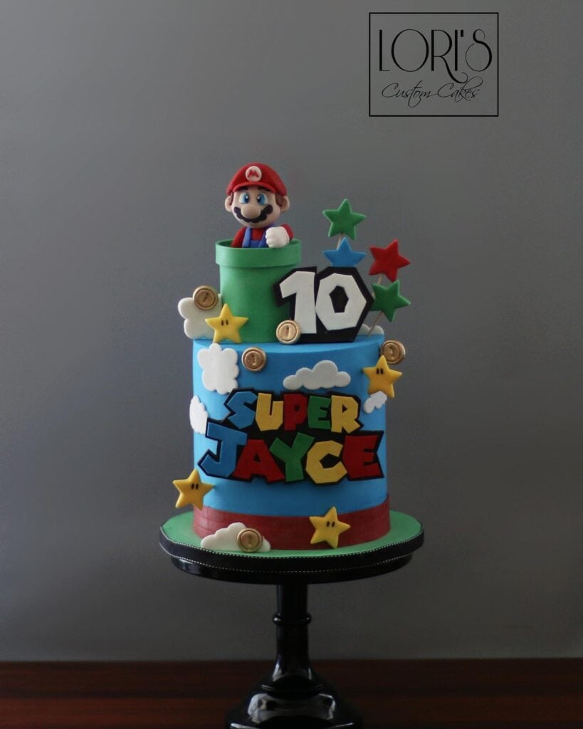A colorful Super Mario-themed birthday cake with Mario figurine, stars, clouds, and the number 10, p.