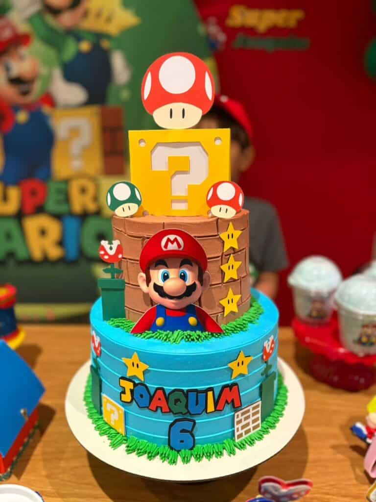 A colorful Super Mario-themed birthday cake with Mario figurine, question blocks, and mushroom decor.