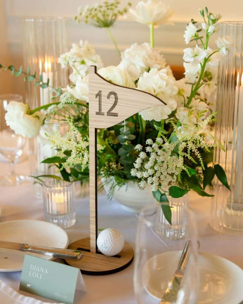 A lush white floral centerpiece with a wooden table number 12 flag for wedding decor.