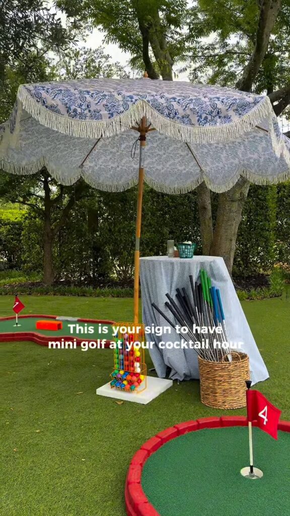 A colorful mini golf setup with umbrella, sign, and accessories on a grassy outdoor area.