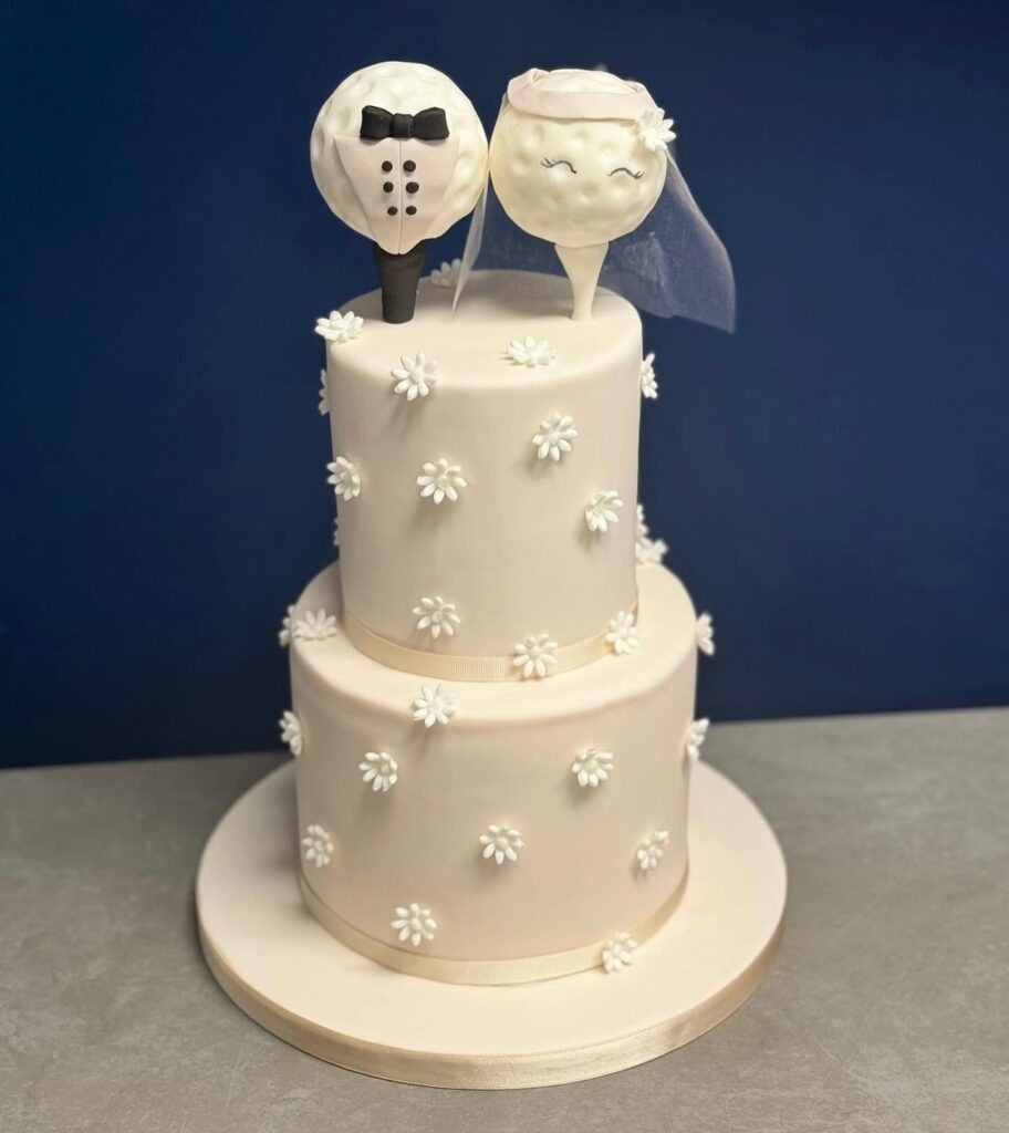 A wedding cake with a tuxedo and bride face topper, decorated with white flowers on a white base.