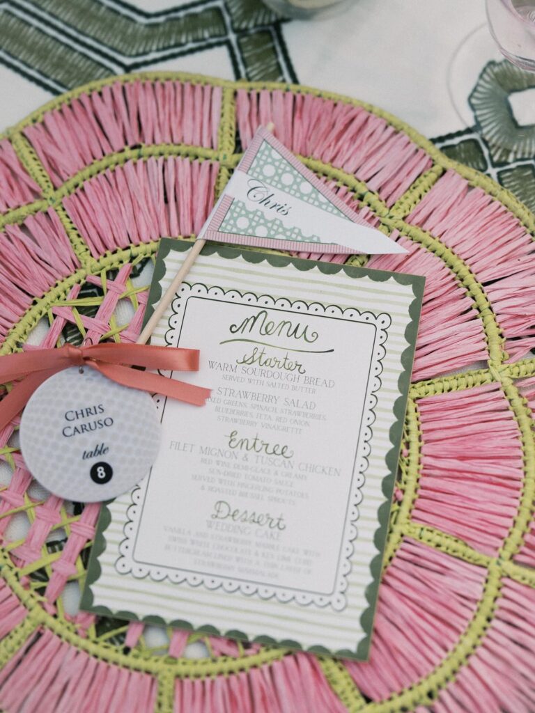 A colorful wedding menu card on a pink and green woven placemat with a pink ribbon and name tag.