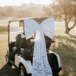 A white golf cart decorated with a large white bow and "Just Married" ribbon on a sunny golf course.