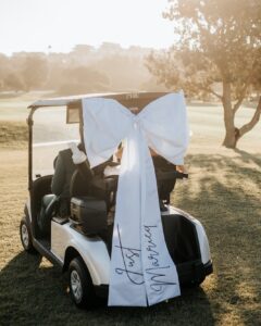 A white golf cart decorated with a large white bow and "Just Married" ribbon on a sunny golf course.