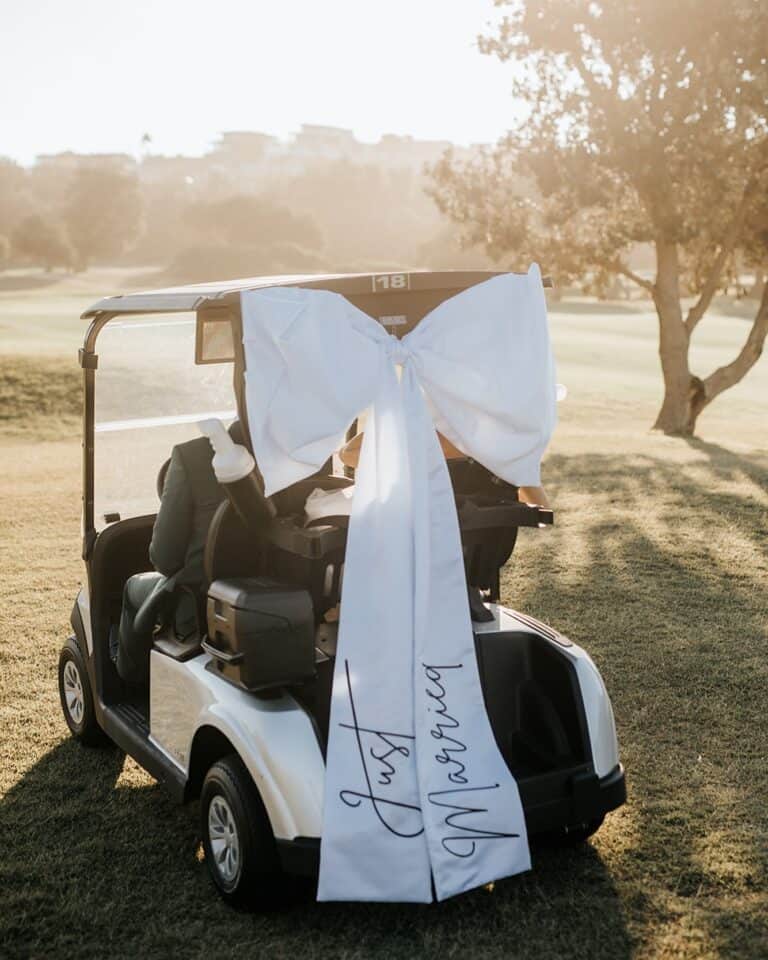 A white golf cart decorated with a large white bow and "Just Married" ribbon on a sunny golf course.