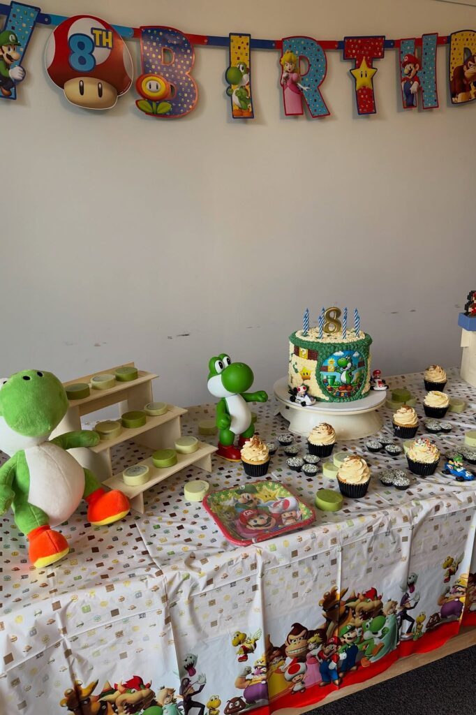 Yoshi-themed birthday party table with cake, cupcakes, and Yoshi plush toys, colorful decorations.