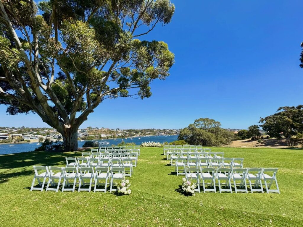 A lush outdoor wedding setup with white chairs and floral arrangements by the water.