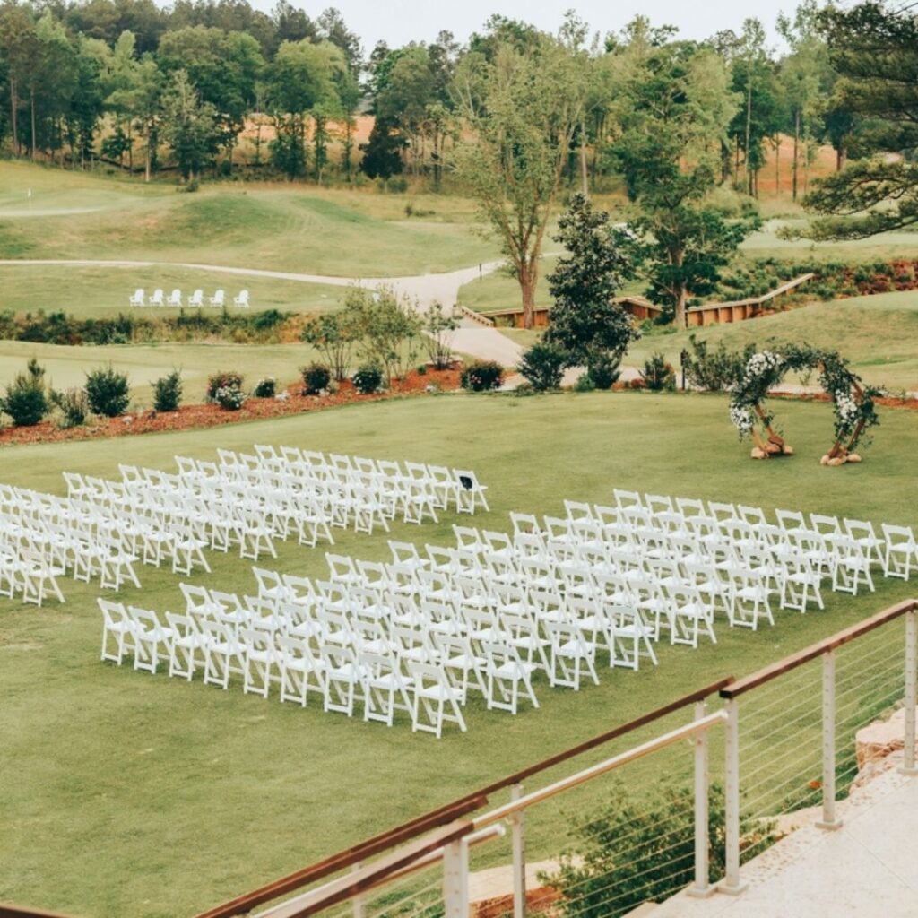 Outdoor wedding ceremony setup with white chairs and floral arch on green lawn.