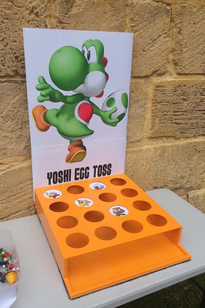 Yoshi egg toss game at a birthday party with Yoshi character backdrop and orange game box.