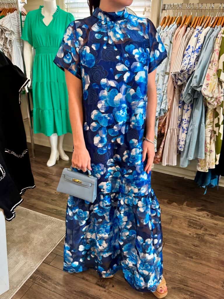 A woman wearing a blue floral maxi dress and holding a small gray handbag in a boutique clothing sto.