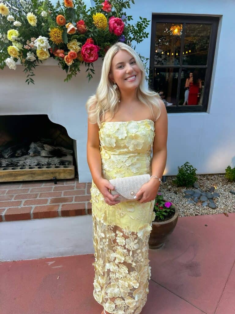 Elegant woman in yellow floral dress holding clutch at outdoor event, beautiful floral decor.