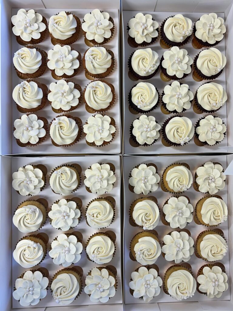 Assorted wedding cupcakes with white floral and swirl frosting designs in a display box.