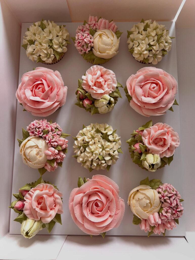 Assorted wedding cupcakes with pink roses and white floral decorations in a box.