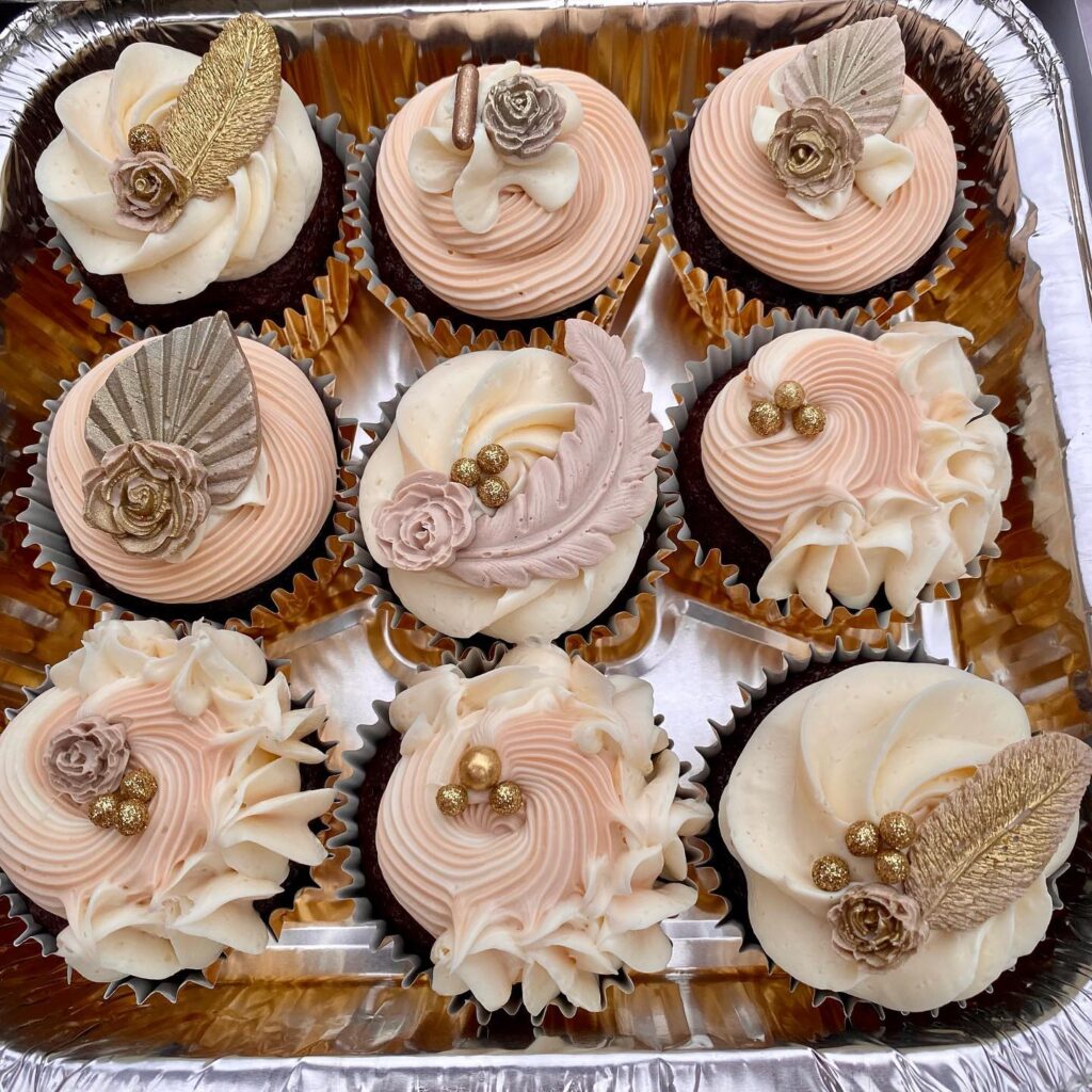 Elegant wedding cupcakes with gold accents, roses, and decorative leaves in a tray.