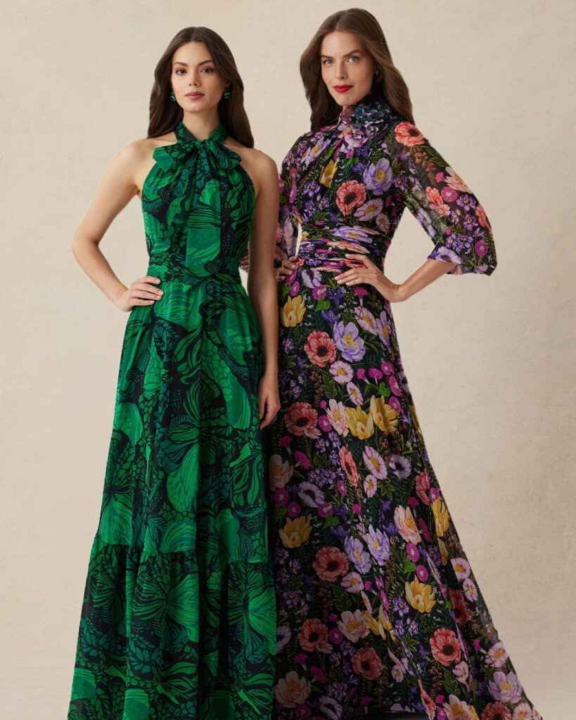A woman in a green tropical print maxi dress and a woman in a floral long-sleeve dress posing togeth.