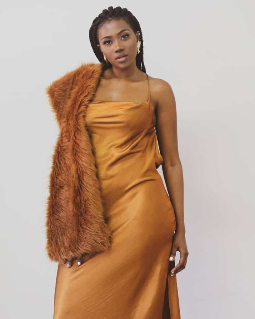 A woman in a satin gold dress with a faux fur coat draped over her shoulder, studio shot.