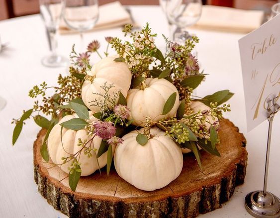 Elegant pumpkin wedding centerpiece with white pumpkins and greenery on a wooden slab.