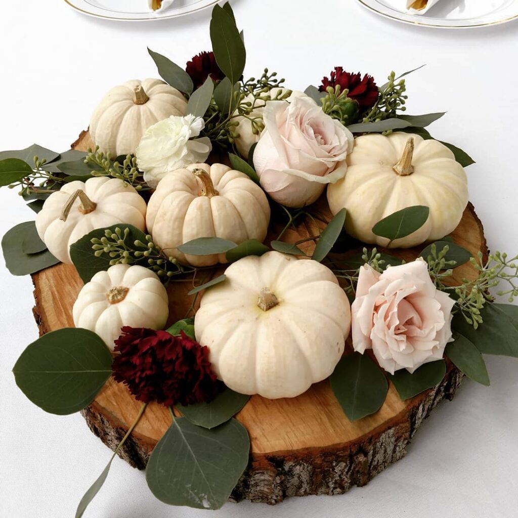 Elegant pumpkin wedding centerpiece with white pumpkins, roses, and greenery on wood slab.