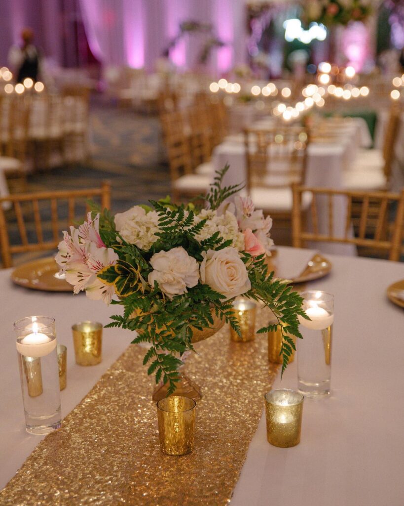 Elegant wedding centerpiece with white roses, greenery, and floating candles on a gold sequin table.