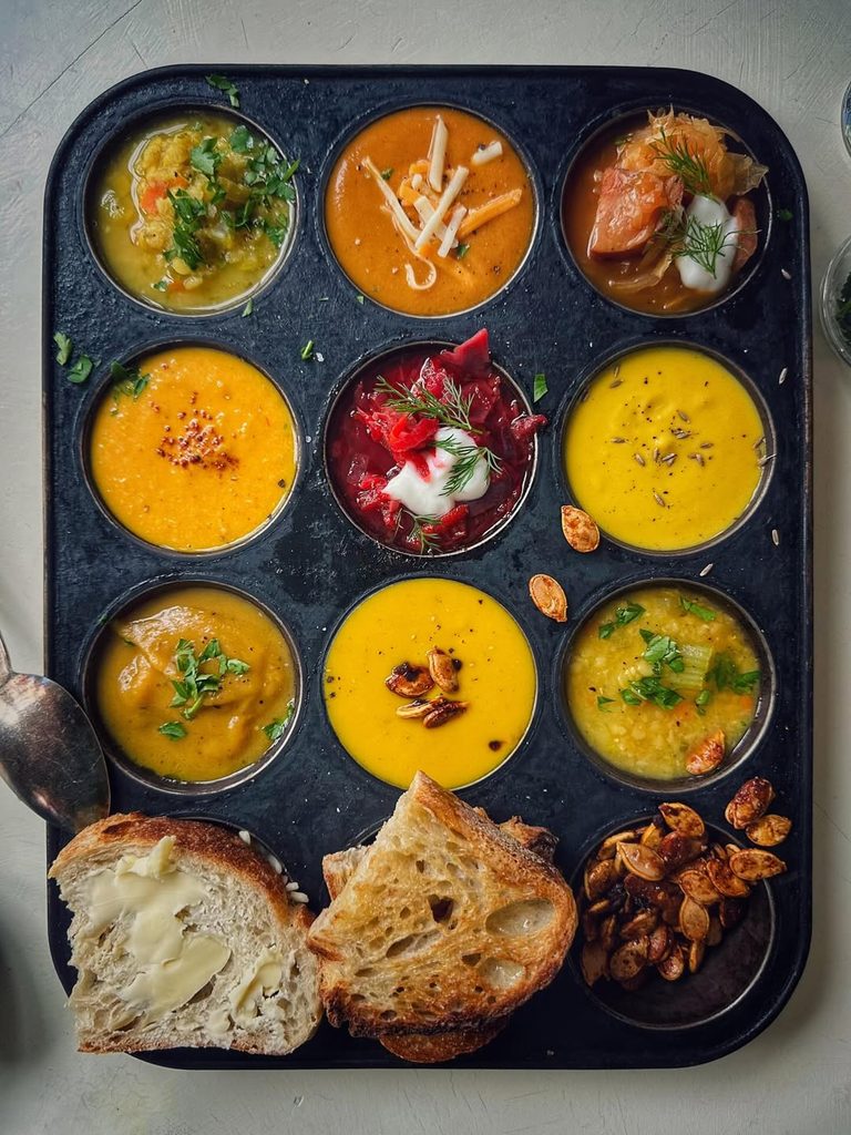 Assorted mini soups in a black tray with bread and toppings for cozy party dining.