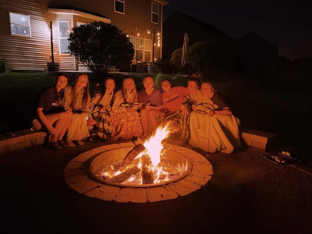 Group of friends sitting around a bonfire at night in a backyard with house lights.