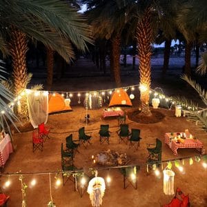 Cozy outdoor bonfire party setup with tents, string lights, and camping chairs among palm trees.