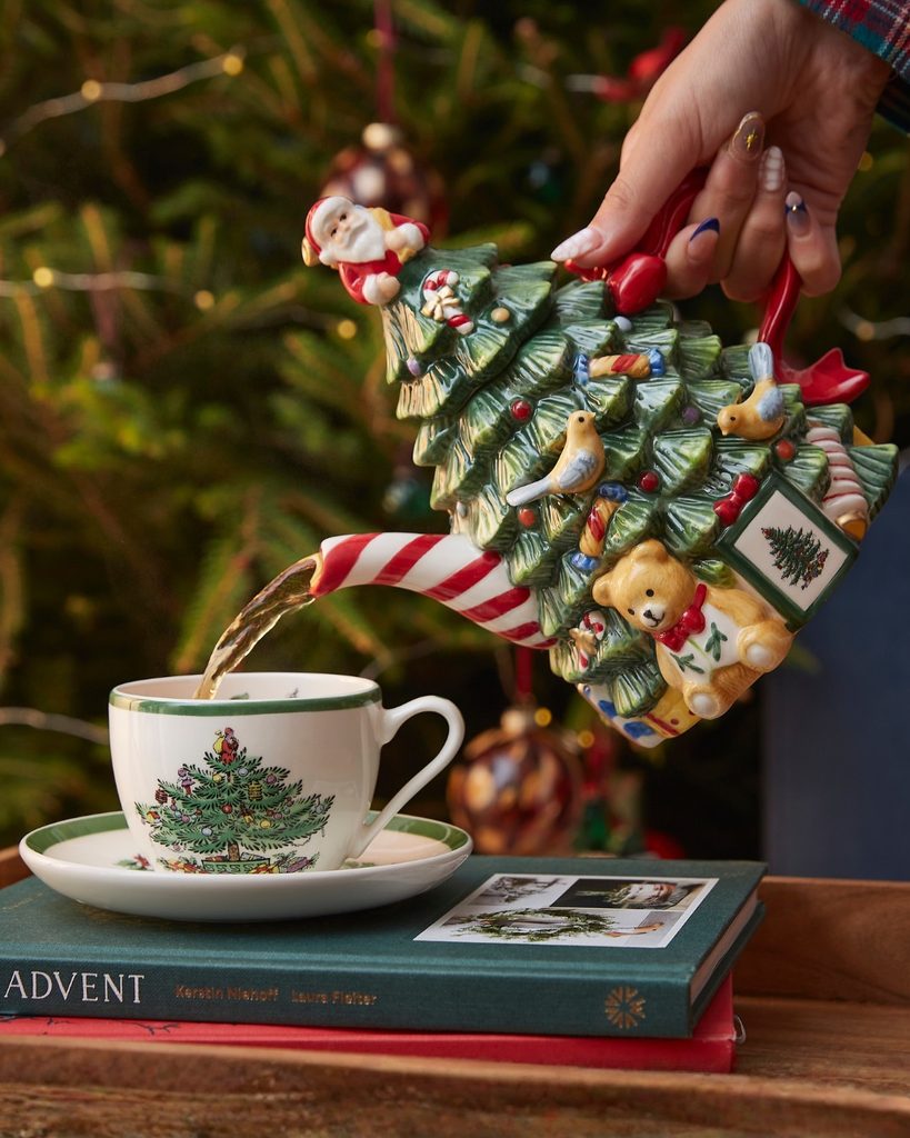 Person pouring tea from a Christmas tree-shaped teapot into a holiday-themed cup.